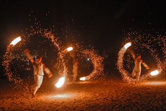 Performers engage in a fire dance, skillfully twirling large fiery torches in circular motions, creating captivating patterns of light against a dark backdrop. The bright flames and flying sparks illuminate their movements as an audience watches the mesmerizing display.