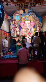 A large and ornate statue of Lord Ganesha, adorned with vibrant colors and intricate patterns, takes center stage. People are gathered around, some standing and some kneeling, suggesting a religious or festive gathering. The backdrop is lavishly decorated with traditional fabrics and hanging garlands.
