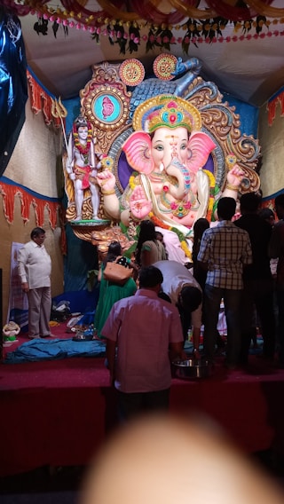 A large and ornate statue of Lord Ganesha, adorned with vibrant colors and intricate patterns, takes center stage. People are gathered around, some standing and some kneeling, suggesting a religious or festive gathering. The backdrop is lavishly decorated with traditional fabrics and hanging garlands.