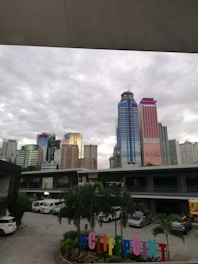 In the foreground, a colorful hashtag sign reads #CLIFFPOINT, while palm trees and various vehicles are visible in the parking area of a building complex. The background features a cityscape with several high-rise buildings, some with brightly lit neon accents, set against a cloudy sky.