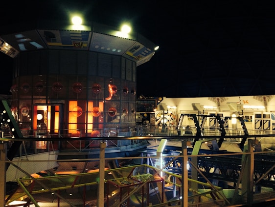 A futuristic, sci-fi interior setting with a distinctive circular structure featuring illuminated panels and a grid of circular designs. The scene includes metallic railings and complex machinery, suggesting a high-tech environment. The lighting is predominantly dim with several warm, glowing lights adding to the ambiance.