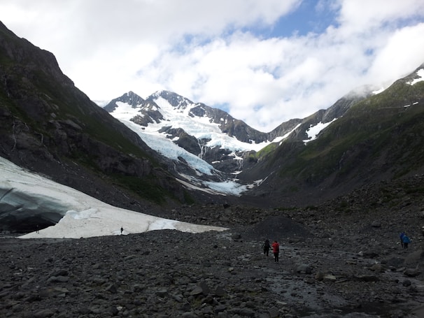 A mountainous landscape with snow-capped peaks and a glacier nestled between the rocky inclines. The foreground comprises rocky terrain with sparse vegetation, and a few people are visible walking along the path. Clouds partially obscure the sky, adding depth to the scene.