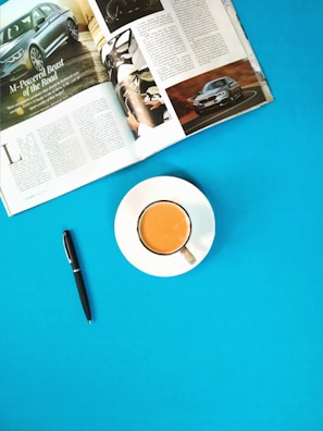 A magazine opened to a page featuring an article about cars is placed on a bright blue surface. Next to it, a cup filled with tea or coffee sits on a white saucer. A black pen is also resting on the same surface, adding to the neat organization.
