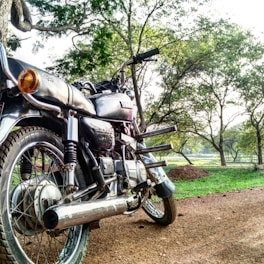 A motorcycle is parked on a dirt path surrounded by a lush, green landscape. The bike appears to be well-maintained, with shiny chrome parts reflecting the sunlight. Tall trees with dense foliage are visible in the background, casting dappled shadows on the ground. The scenery suggests a peaceful, natural setting, possibly in a park or countryside.