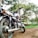 A motorcycle is parked on a dirt path surrounded by a lush, green landscape. The bike appears to be well-maintained, with shiny chrome parts reflecting the sunlight. Tall trees with dense foliage are visible in the background, casting dappled shadows on the ground. The scenery suggests a peaceful, natural setting, possibly in a park or countryside.