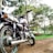 A motorcycle is parked on a dirt path surrounded by a lush, green landscape. The bike appears to be well-maintained, with shiny chrome parts reflecting the sunlight. Tall trees with dense foliage are visible in the background, casting dappled shadows on the ground. The scenery suggests a peaceful, natural setting, possibly in a park or countryside.