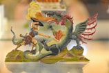 A detailed miniature figure depicting a dramatic scene of a warrior battling a fierce dragon. The warrior, wearing ancient-style armor and a helmet, is poised to attack with a sword. The dragon, with vibrant green and red scales, is shown breathing fire towards the warrior. The base is designed to resemble rocky terrain.