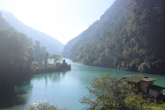 A serene river winds through a lush, mountainous landscape. Dense green forests cover the hills on either side, creating a sense of seclusion and tranquility. A small house with a sloping roof sits on the right bank of the river, partially obscured by foliage. The sky is bright with mist, filtering the sunlight over the scene.