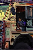 A colorfully decorated truck features intricate patterns and vibrant designs. The open cabin door reveals a person reclining inside, wearing a patterned garment and flip-flops. The truck displays writings and adornments with bold colors, creating a lively and detailed appearance.