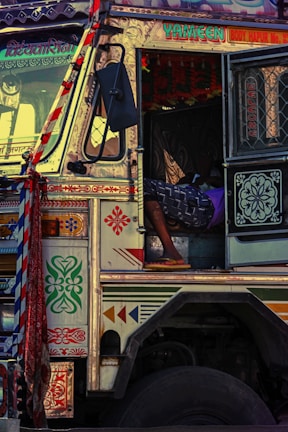 A colorfully decorated truck features intricate patterns and vibrant designs. The open cabin door reveals a person reclining inside, wearing a patterned garment and flip-flops. The truck displays writings and adornments with bold colors, creating a lively and detailed appearance.