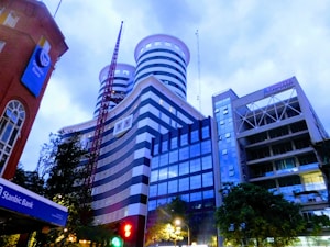 A cityscape featuring a group of modern high-rise buildings with unique cylindrical and striped designs. The scene is captured during dusk or early evening, indicated by the bluish tint in the sky and illuminated streetlights. A red crane is visible in the foreground, and signs for a bank and other businesses are apparent on the buildings.