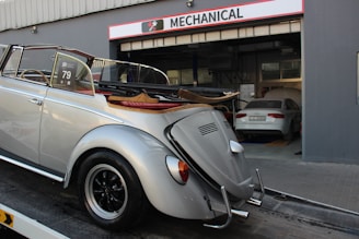 A classic silver convertible car is parked on a trailer in front of a garage with the sign 'Mechanical' above the entrance. The car has vintage styling and a soft top that is folded down. Inside the garage, there is another car parked, which appears to be a modern sedan. The environment suggests a car service or repair facility.