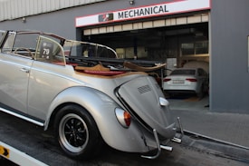 A classic silver convertible car is parked on a trailer in front of a garage with the sign 'Mechanical' above the entrance. The car has vintage styling and a soft top that is folded down. Inside the garage, there is another car parked, which appears to be a modern sedan. The environment suggests a car service or repair facility.