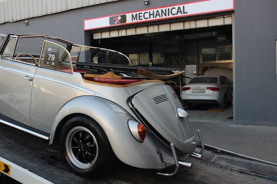 A classic silver convertible car is parked on a trailer in front of a garage with the sign 'Mechanical' above the entrance. The car has vintage styling and a soft top that is folded down. Inside the garage, there is another car parked, which appears to be a modern sedan. The environment suggests a car service or repair facility.