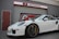 A sleek white sports car is parked in front of a garage with a sign that reads 'MECHANICAL'. The car features intricate design details and polished alloy wheels. The garage has a metal roller shutter with a logo that includes gears and wrenches.