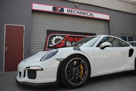 A sleek white sports car is parked in front of a garage with a sign that reads 'MECHANICAL'. The car features intricate design details and polished alloy wheels. The garage has a metal roller shutter with a logo that includes gears and wrenches.
