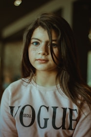 A young girl with long dark hair looks forward, wearing a light pink sweatshirt with the word 'VOGUE' printed on it. The lighting is soft, creating a warm atmosphere.