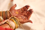 Intricately decorated hands with henna designs are holding small grains. The wrists are adorned with colorful bangles, predominantly in shades of gold, green, and orange, adding a festive and traditional touch.