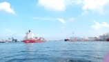 A cargo ship with colorful containers navigates through a body of water under a clear blue sky. Industrial cranes and dock facilities are visible on the left side, while additional ships and port structures are in the background.