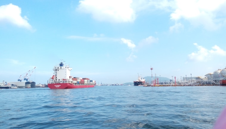 A cargo ship with colorful containers navigates through a body of water under a clear blue sky. Industrial cranes and dock facilities are visible on the left side, while additional ships and port structures are in the background.