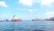 A cargo ship with colorful containers navigates through a body of water under a clear blue sky. Industrial cranes and dock facilities are visible on the left side, while additional ships and port structures are in the background.
