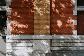 A section of a brick wall in alternating patterns of red and light brown bricks. Gray stone blocks frame the bricks and are also part of the lower wall. Shadows cast by foliage create a dappled effect on the wall surface.