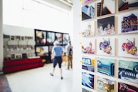 A gallery display featuring vibrant, colorful artworks on a wall. The art includes abstract, multicolored depictions of buildings, animals, and cars. In the blurred background, two people are observing additional artworks that are hung on another wall.