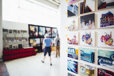 A gallery display featuring vibrant, colorful artworks on a wall. The art includes abstract, multicolored depictions of buildings, animals, and cars. In the blurred background, two people are observing additional artworks that are hung on another wall.