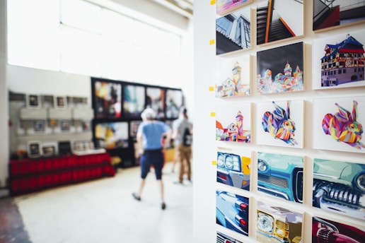 A gallery display featuring vibrant, colorful artworks on a wall. The art includes abstract, multicolored depictions of buildings, animals, and cars. In the blurred background, two people are observing additional artworks that are hung on another wall.