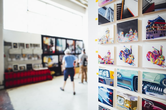 A gallery display featuring vibrant, colorful artworks on a wall. The art includes abstract, multicolored depictions of buildings, animals, and cars. In the blurred background, two people are observing additional artworks that are hung on another wall.