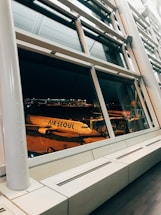A large glass window in an airport terminal provides a view of an airplane with the 'Air Seoul' logo, which is parked at the gate during nighttime. The scene is illuminated by artificial lights, and the interior features modern architectural elements like metallic beams and clean lines.