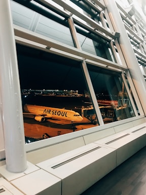 A large glass window in an airport terminal provides a view of an airplane with the 'Air Seoul' logo, which is parked at the gate during nighttime. The scene is illuminated by artificial lights, and the interior features modern architectural elements like metallic beams and clean lines.