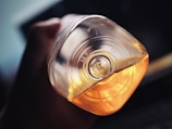 A close-up view of a hand holding an inverted plastic bottle with an embossed recycling symbol on the bottom. The bottle contains a yellow-orange liquid, and the lighting highlights the transparency and reflections on the bottle, adding depth to the image.