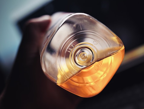 A close-up view of a hand holding an inverted plastic bottle with an embossed recycling symbol on the bottom. The bottle contains a yellow-orange liquid, and the lighting highlights the transparency and reflections on the bottle, adding depth to the image.