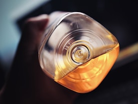 A close-up view of a hand holding an inverted plastic bottle with an embossed recycling symbol on the bottom. The bottle contains a yellow-orange liquid, and the lighting highlights the transparency and reflections on the bottle, adding depth to the image.