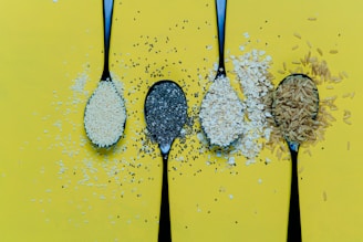Four black spoons are lined up against a bright yellow background. Each spoon contains different types of grains or seeds: sesame seeds, chia seeds, oats, and rice. The various grains spill over the edges of the spoons, creating a textured pattern on the background.