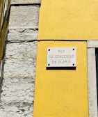 A street sign is mounted on a vibrant yellow wall next to a section of exposed stonework on a building. The sign reads 'Rua da Conceição da Glória' and is attached to the wall with visible screws.