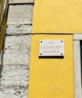 A street sign is mounted on a vibrant yellow wall next to a section of exposed stonework on a building. The sign reads 'Rua da Conceição da Glória' and is attached to the wall with visible screws.
