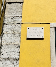 A street sign is mounted on a vibrant yellow wall next to a section of exposed stonework on a building. The sign reads 'Rua da Conceição da Glória' and is attached to the wall with visible screws.