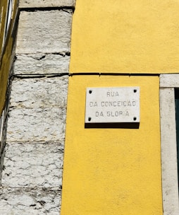A street sign is mounted on a vibrant yellow wall next to a section of exposed stonework on a building. The sign reads 'Rua da Conceição da Glória' and is attached to the wall with visible screws.