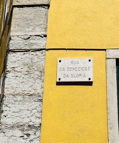 A street sign is mounted on a vibrant yellow wall next to a section of exposed stonework on a building. The sign reads 'Rua da Conceição da Glória' and is attached to the wall with visible screws.