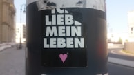 A sticker is attached to a pole with the words 'ICH LIEBE MEIN LEBEN' in bold white letters, accompanied by a small pink heart graphic underneath. The background is an urban street scene with blurred buildings and a curved roadway in the distance.