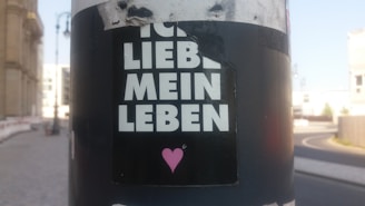 A sticker is attached to a pole with the words 'ICH LIEBE MEIN LEBEN' in bold white letters, accompanied by a small pink heart graphic underneath. The background is an urban street scene with blurred buildings and a curved roadway in the distance.