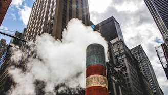 A city street scene with tall skyscrapers against a partly cloudy sky. A large plume of steam is rising from a striped steam pipe in the center. The surrounding buildings have various architectural styles, and a street sign reading 'Dey St' is visible.