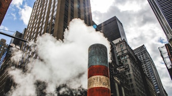 A city street scene with tall skyscrapers against a partly cloudy sky. A large plume of steam is rising from a striped steam pipe in the center. The surrounding buildings have various architectural styles, and a street sign reading 'Dey St' is visible.