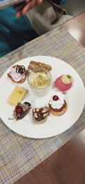 A variety of small, intricate desserts are arranged on a white plate. There are selections including a small tart with pink cream and chocolate topping, a glass cup containing a creamy dessert with nuts, a round pink dessert with cream and a small green garnish, a square piece of creamy yellow dessert with a chocolate decoration, a chocolate mousse dessert with a cherry, and two mini cakes garnished with whipped cream and nuts. The plate rests on a colorful fabric placemat, and a hand holding a phone is partially visible in the top left corner.