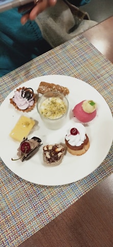 A variety of small, intricate desserts are arranged on a white plate. There are selections including a small tart with pink cream and chocolate topping, a glass cup containing a creamy dessert with nuts, a round pink dessert with cream and a small green garnish, a square piece of creamy yellow dessert with a chocolate decoration, a chocolate mousse dessert with a cherry, and two mini cakes garnished with whipped cream and nuts. The plate rests on a colorful fabric placemat, and a hand holding a phone is partially visible in the top left corner.