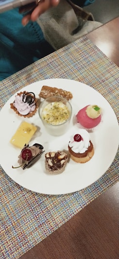 A variety of small, intricate desserts are arranged on a white plate. There are selections including a small tart with pink cream and chocolate topping, a glass cup containing a creamy dessert with nuts, a round pink dessert with cream and a small green garnish, a square piece of creamy yellow dessert with a chocolate decoration, a chocolate mousse dessert with a cherry, and two mini cakes garnished with whipped cream and nuts. The plate rests on a colorful fabric placemat, and a hand holding a phone is partially visible in the top left corner.