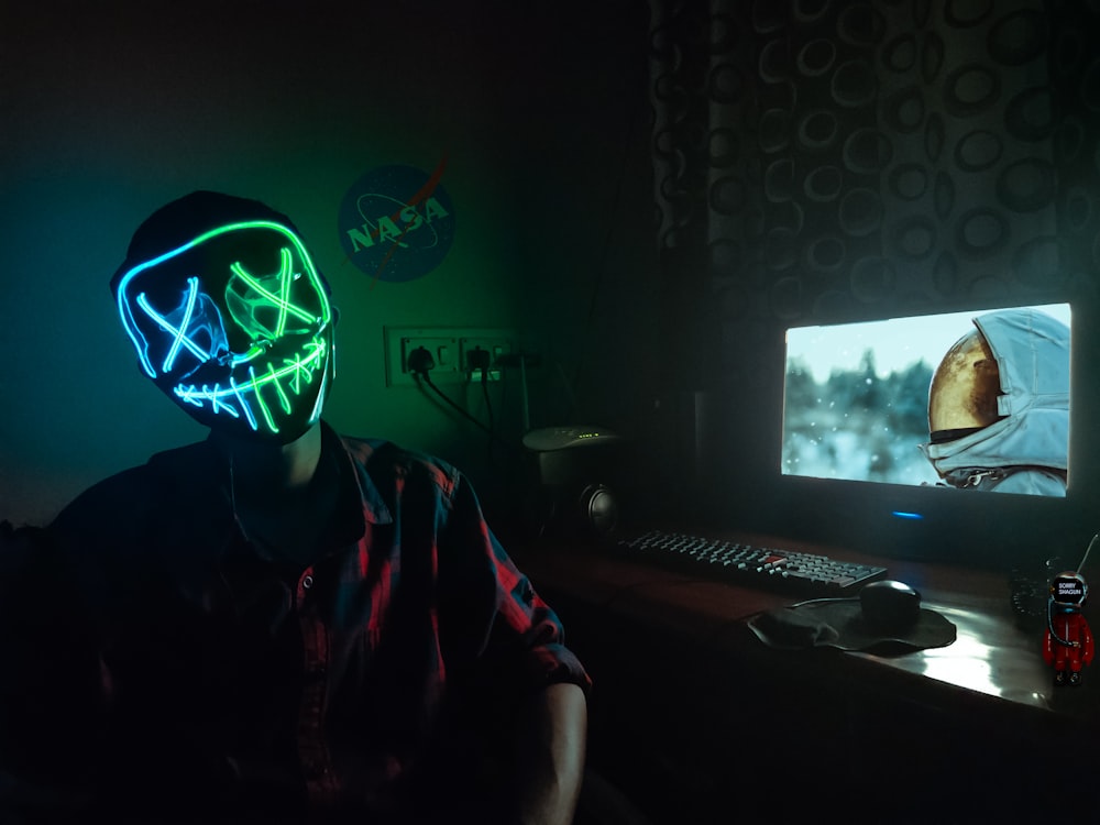 person in blue and green LEd mask sitting in front of turned-on computer monitor