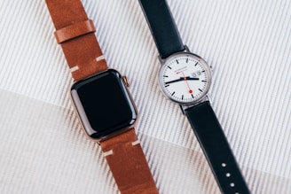 A modern digital smartwatch with a brown leather band is placed next to a classic analog wristwatch with a black leather band. The digital watch has a sleek, black rectangular face, while the analog watch features a white face with a date display and red accents on the hands.